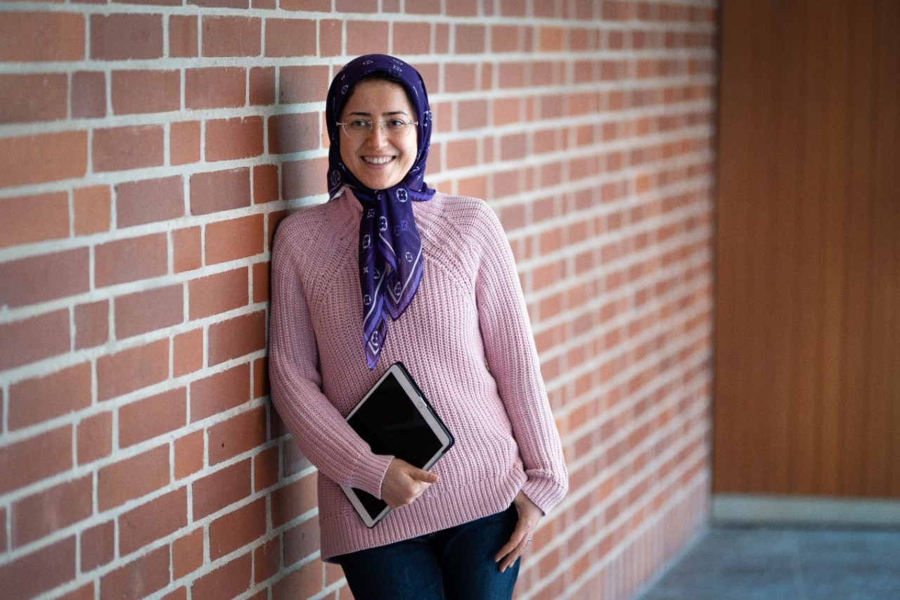 A woman in a pink knitted sweater and purple headscarf leans against a brick wall. She is smiling and holding an iPad.