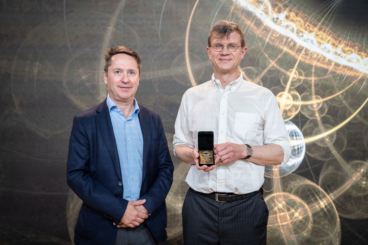 Two male scientists standing next to eachother, one holding a golden medal. 