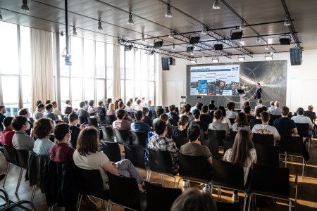 An audience in a brightly-lit lecture hall watches a speaker onstage.