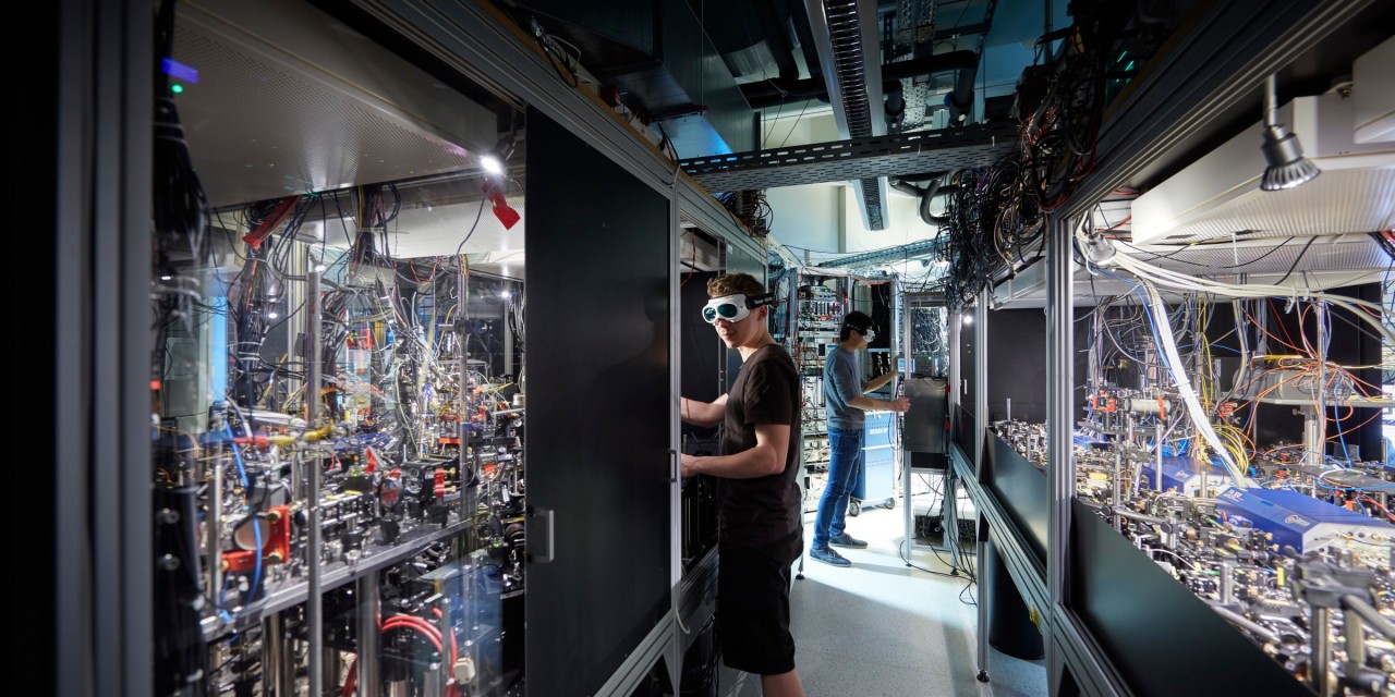 Benches with lasers, fiber optics, lenses, reflection mirrors, and vacuum chambers in an quantum optics laboratory.