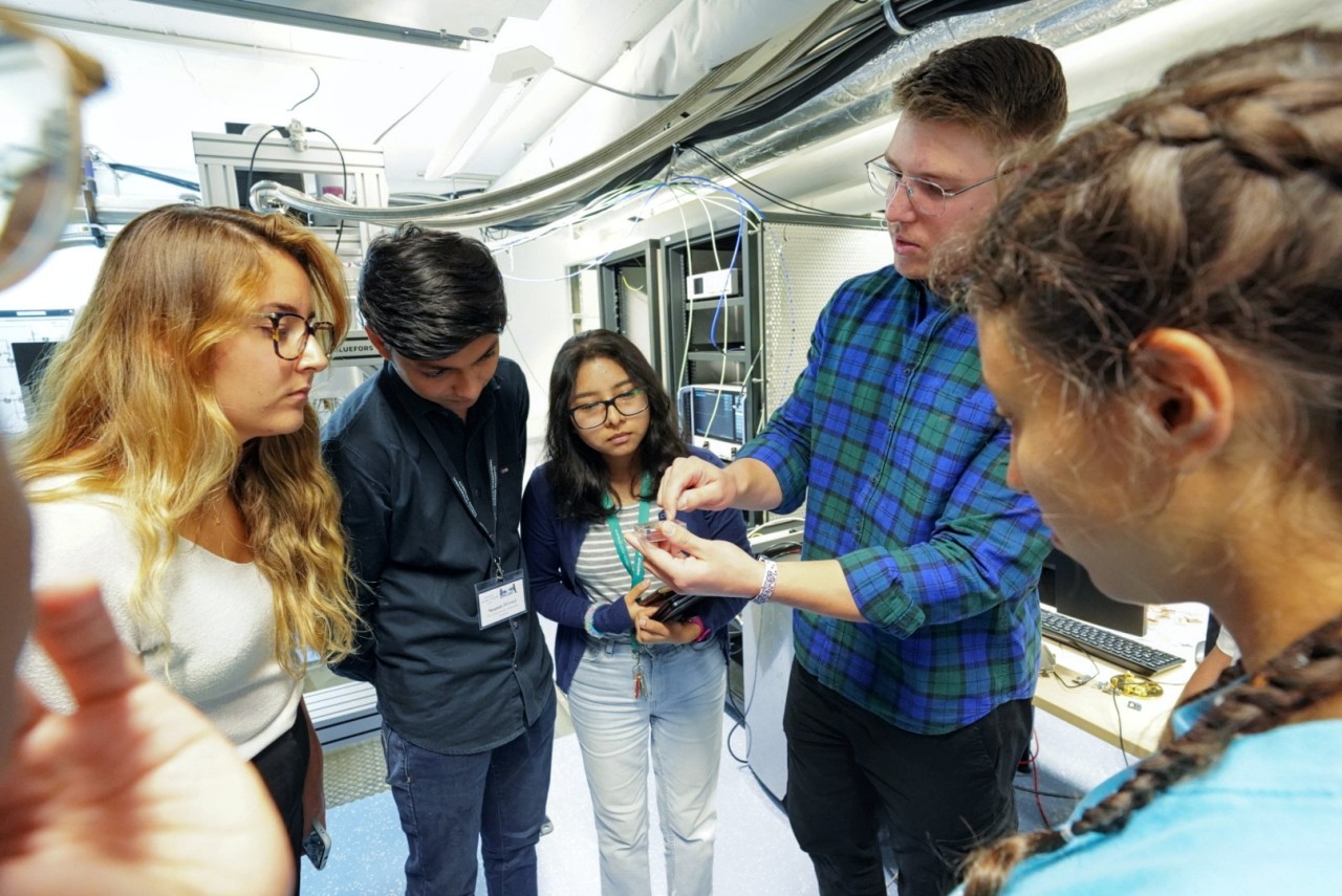 Students gathered around a scientists, who is showing a small piece the researchers are currently working on. 